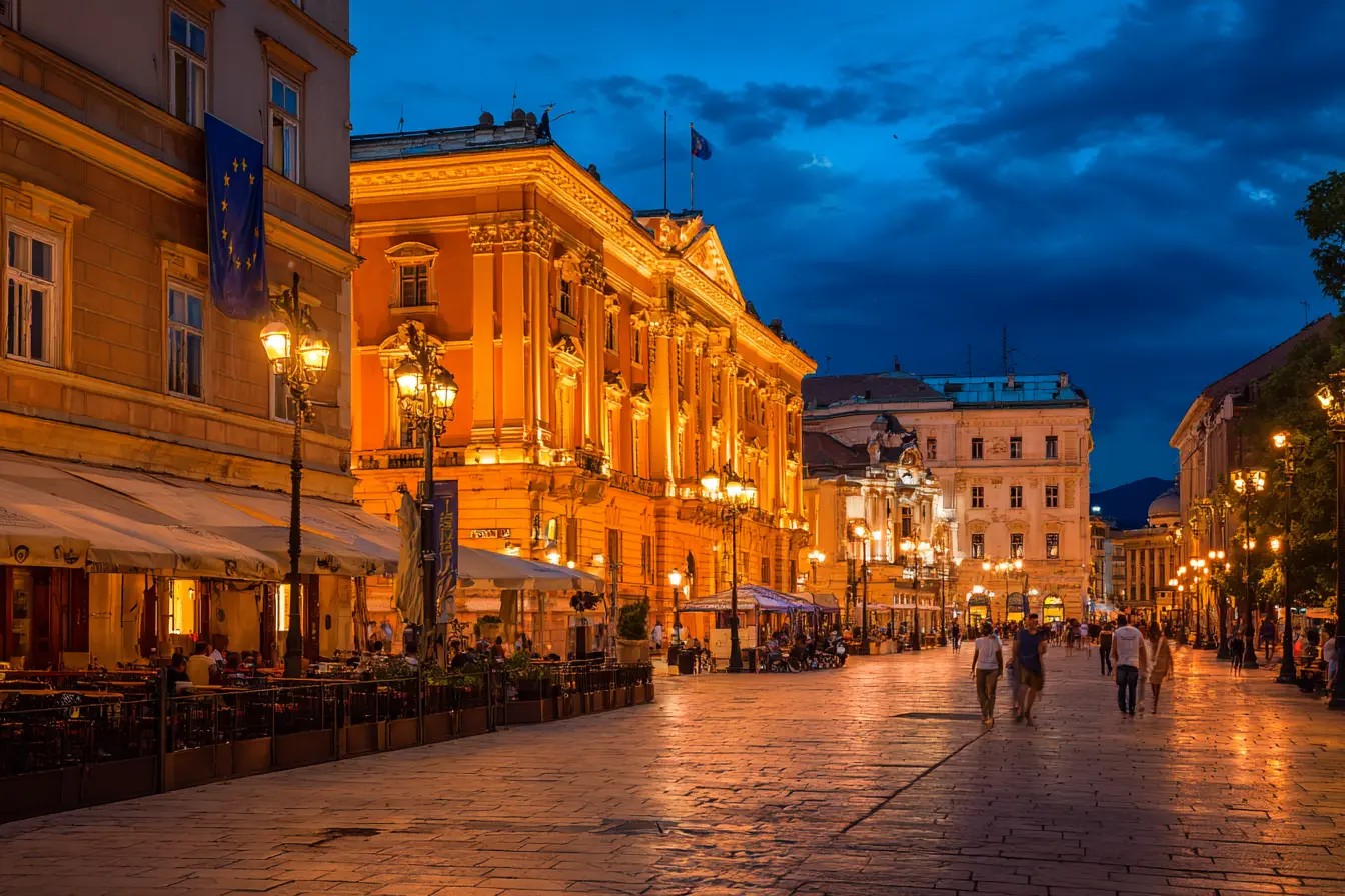 La piazza centrale di una capitale dell’Europa orientale di sera: la facciata illuminata di un palazzo di governo, una bandiera dell’Unione ferma sul pennone, gente ai tavolini dei caffè, riflessi sul selciato umido.