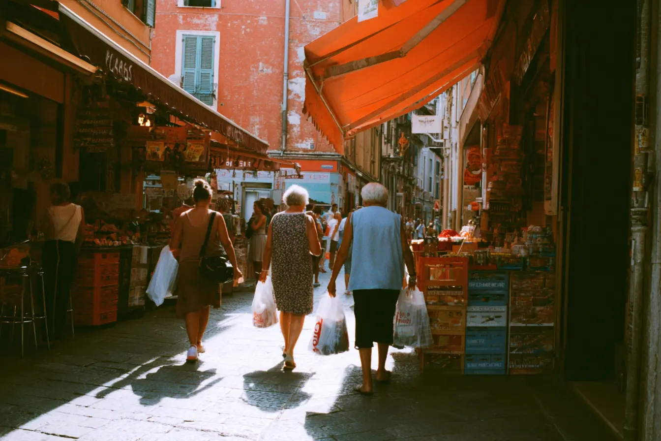 Scatto rubato in una via commerciale di una città italiana nel tardo pomeriggio: gente con le buste della spesa, un’edicola, un bar con i tavolini fuori, tende da sole.